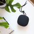 Black portable wireless speaker with a rounded square design and carrying strap on a white desk beside green plants and a book by sackit