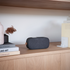 Black sackit Go 300 Care portable Bluetooth speaker displayed on a glass shelf in a modern home interior with green plants and wood paneling