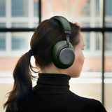 Woman wearing green sackit over ear headphones while looking out a window indoors with soft natural light and a modern city view beyond