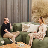 Two people relaxing on a green Soft Sofa Outdoor corner sofa beside a wooden table with cushions and drinks near coastal grasses from sackit