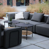 Dark gray modular outdoor corner sofa with cushions and black nesting coffee tables on a paved patio in a landscaped garden designed by sackit