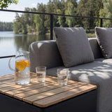 Grey outdoor lounge sofa beside a teak patio storage table with pitcher and glasses overlooking a calm lake in Scandinavian surroundings by sackit