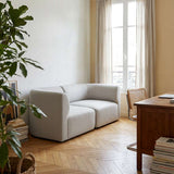 Light gray two seater sofa by sackit in a calm Scandinavian living room with a window plant wooden desk and herringbone floor