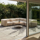 Beige outdoor lounge sofa with a bolster cushion and round coffee table on a modern patio surrounded by lush greenery viewed through open glass doors