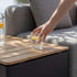 Teak outdoor storage table beside a grey lounge sofa with a hand placing a glass of lemon water on the sunlit tabletop