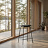 Black leather bar stool with a slim metal frame beside floor to ceiling windows in a modern Nordic interior overlooking a forest lake by sackit