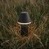 Black cordless outdoor table lamp glowing softly among tall meadow grass for atmospheric Nordic evenings by sackit
