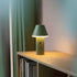 sackit Cone cordless table lamp in green glowing softly on a wooden shelf beside books in a cosy indoor setting
