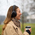 Woman wearing black over ear headphones and holding a takeaway coffee outdoors on a cool day
