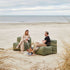 Two people relax on green Soft Sofa Outdoor modules by sackit on a sandy beach beside the sea demonstrating comfortable outdoor lounge furniture