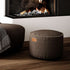 Round brown canvas pouf beside a modern fireplace in a Nordic living room designed by sackit for comfortable indoor lounging
