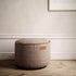 Brown canvas indoor pouf with a rounded shape and decorative stitched edge on a wooden floor in a calm neutral interior by sackit