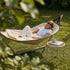 Woman relaxing in a Cobana hammock on a sunny garden lawn beside a small outdoor table with coffee and reading material