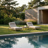 Two sackit outdoor sunbeds with light cushions beside a swimming pool in a landscaped garden with ornamental grasses pine trees and modern architecture