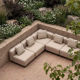 Beige modular outdoor corner sofa with plush cushions on a brick patio surrounded by lush flowers and greenery in a calm Scandinavian garden setting