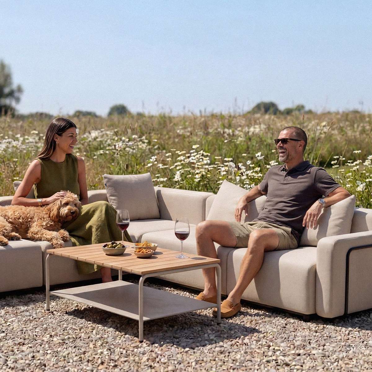 sackit Cobana outdoor corner sofa with two people and a dog relaxing around a wooden coffee table in a sunny meadow