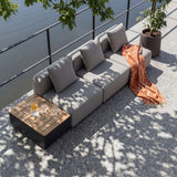 Gray three seat sackit outdoor lounge sofa with cushions and wooden patio storage table on a sunlit terrace beside a glass railing and greenery