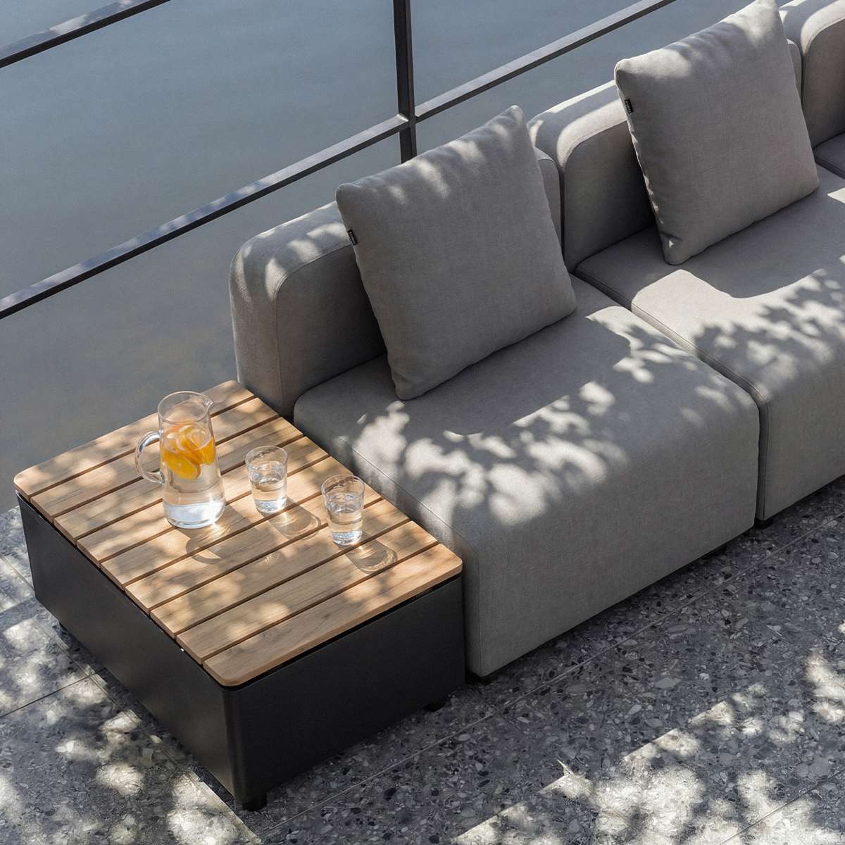 Gray modular outdoor lounge sofa with cushions beside a wooden topped side table and drinks on a sunny terrace designed in Denmark by sackit Europe