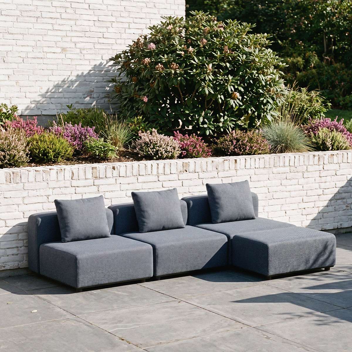 Dark gray three seat modular outdoor lounge sofa with cushions on a paved terrace beside a landscaped garden designed in Denmark by sackit