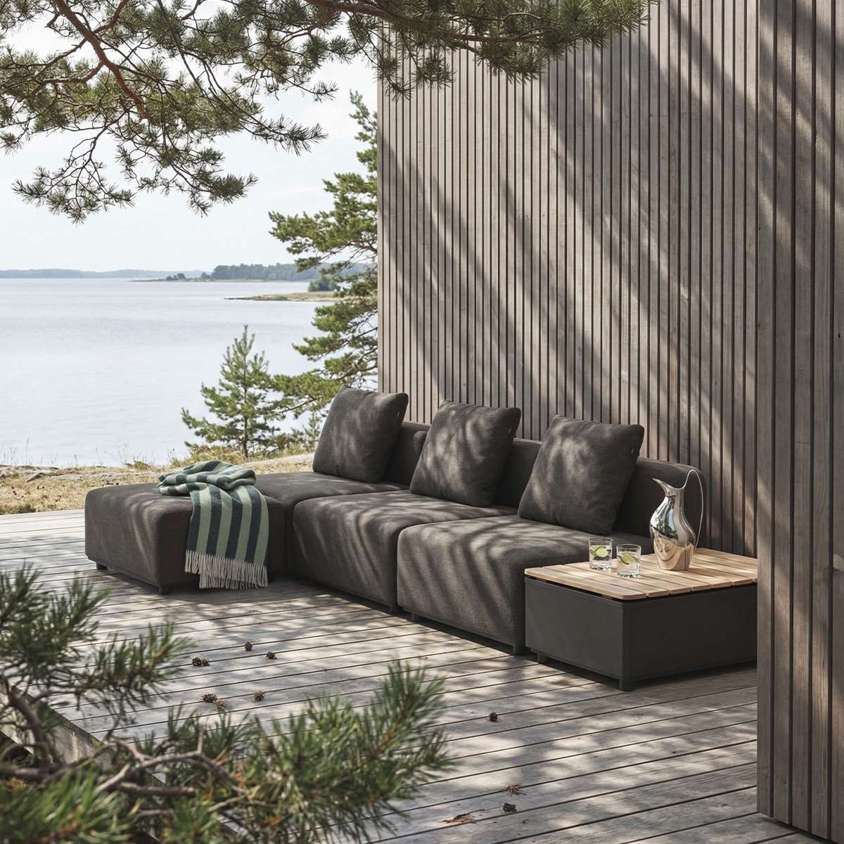 sackit dark modular outdoor lounge sofa with cushions and striped throw on a wooden coastal deck beside the sea and a pine lined Scandinavian cabin