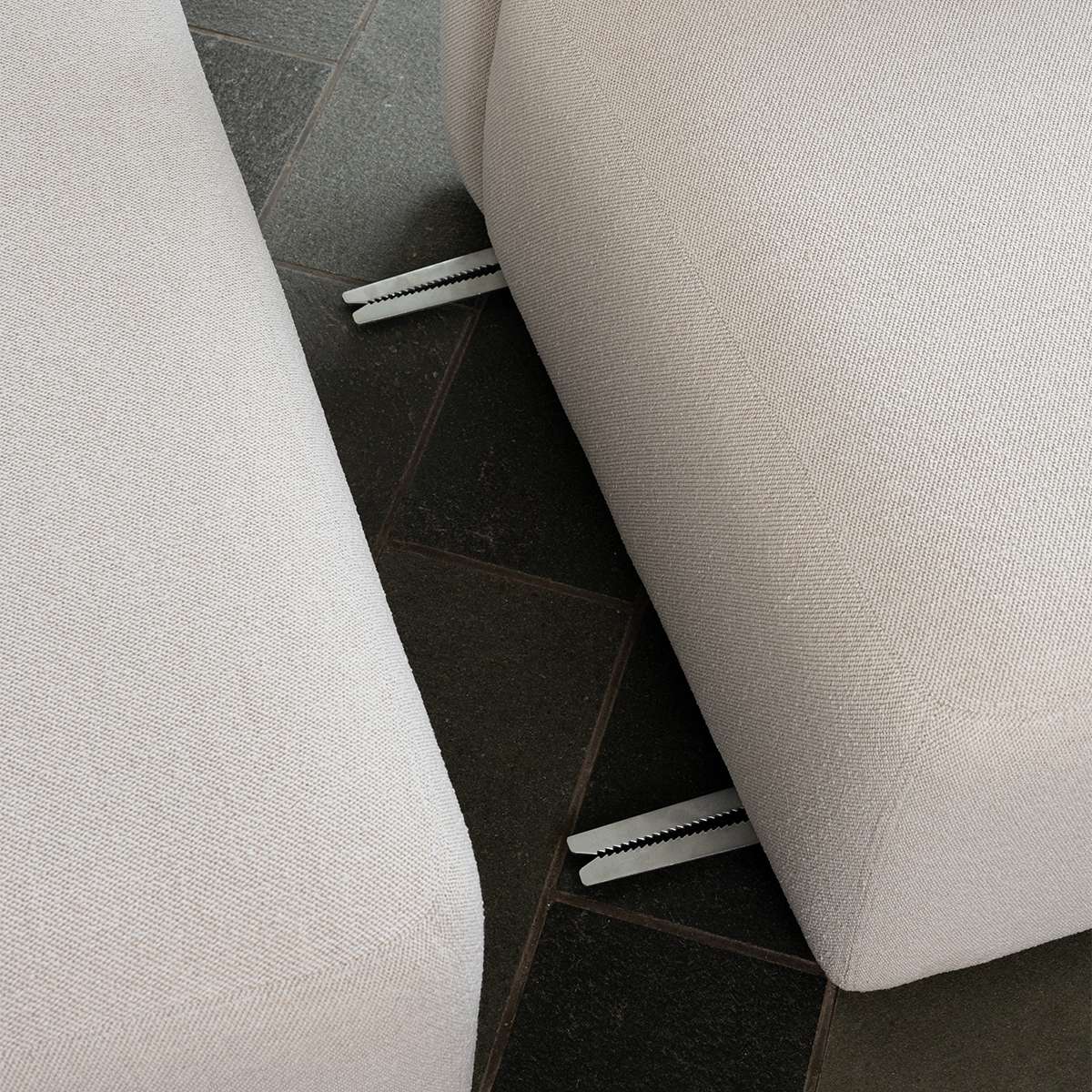 Close up of a light fabric sofa with visible metal joining brackets between cushioned sections on a dark tiled floor from sackit