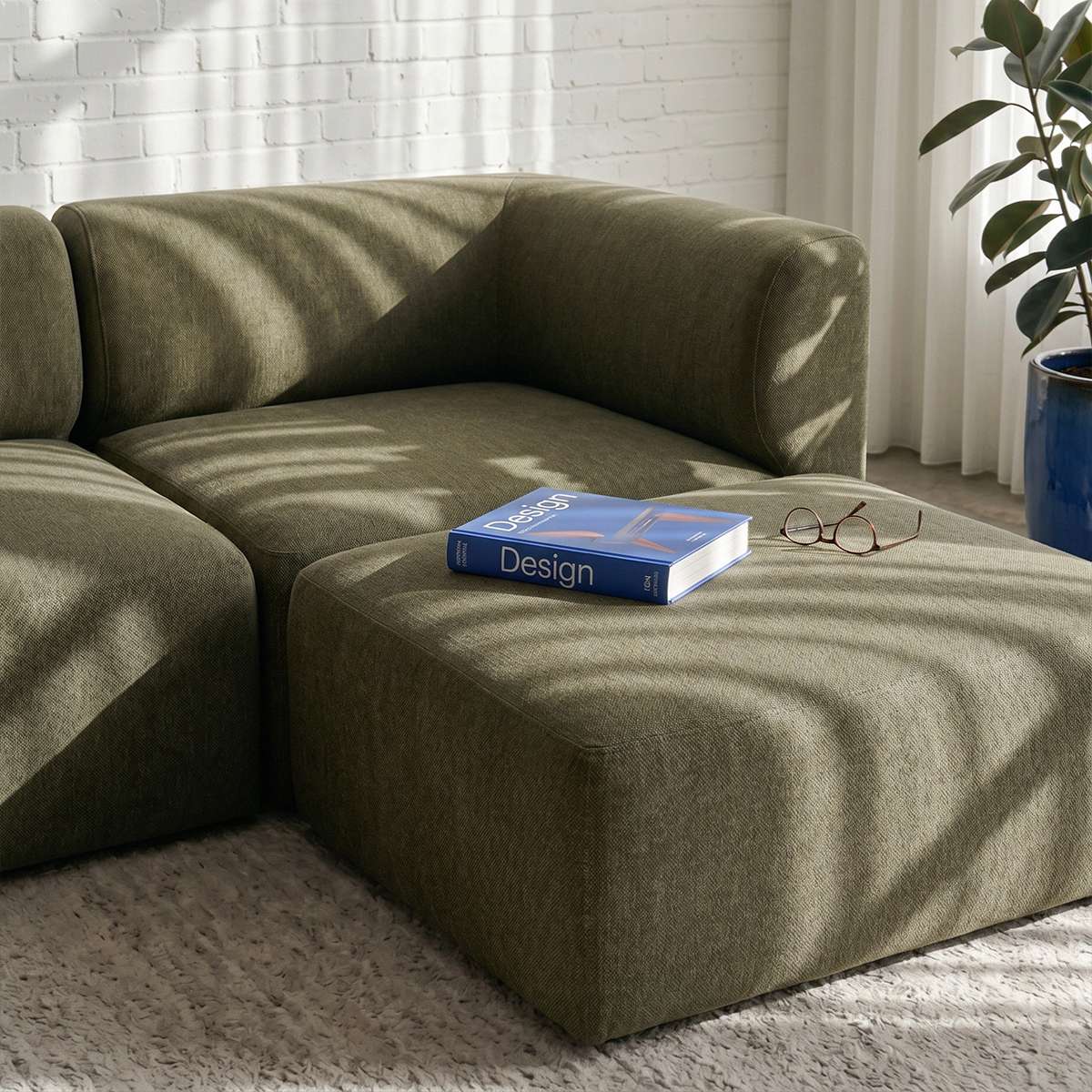 Olive green Fjord sofa with matching pouf in a sunlit Scandinavian living room beside a blue design book and eyeglasses from sackit