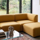 Mustard yellow Fjord sofa with matching pouf in a bright Scandinavian living room beside a glass coffee table and patterned rug