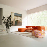 Orange three seat indoor sofa with lounger and patterned ottoman in a bright modern living room with artwork houseplant and large window from sackit