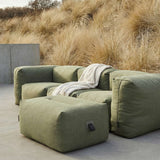 Sage green outdoor pouf beside a matching lounge sofa with a soft throw in a natural Nordic garden setting by sackit