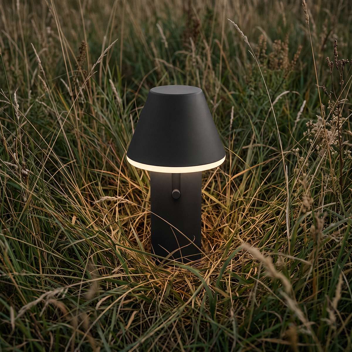 Black cordless outdoor table lamp glowing softly among tall meadow grass for atmospheric Nordic evenings by sackit