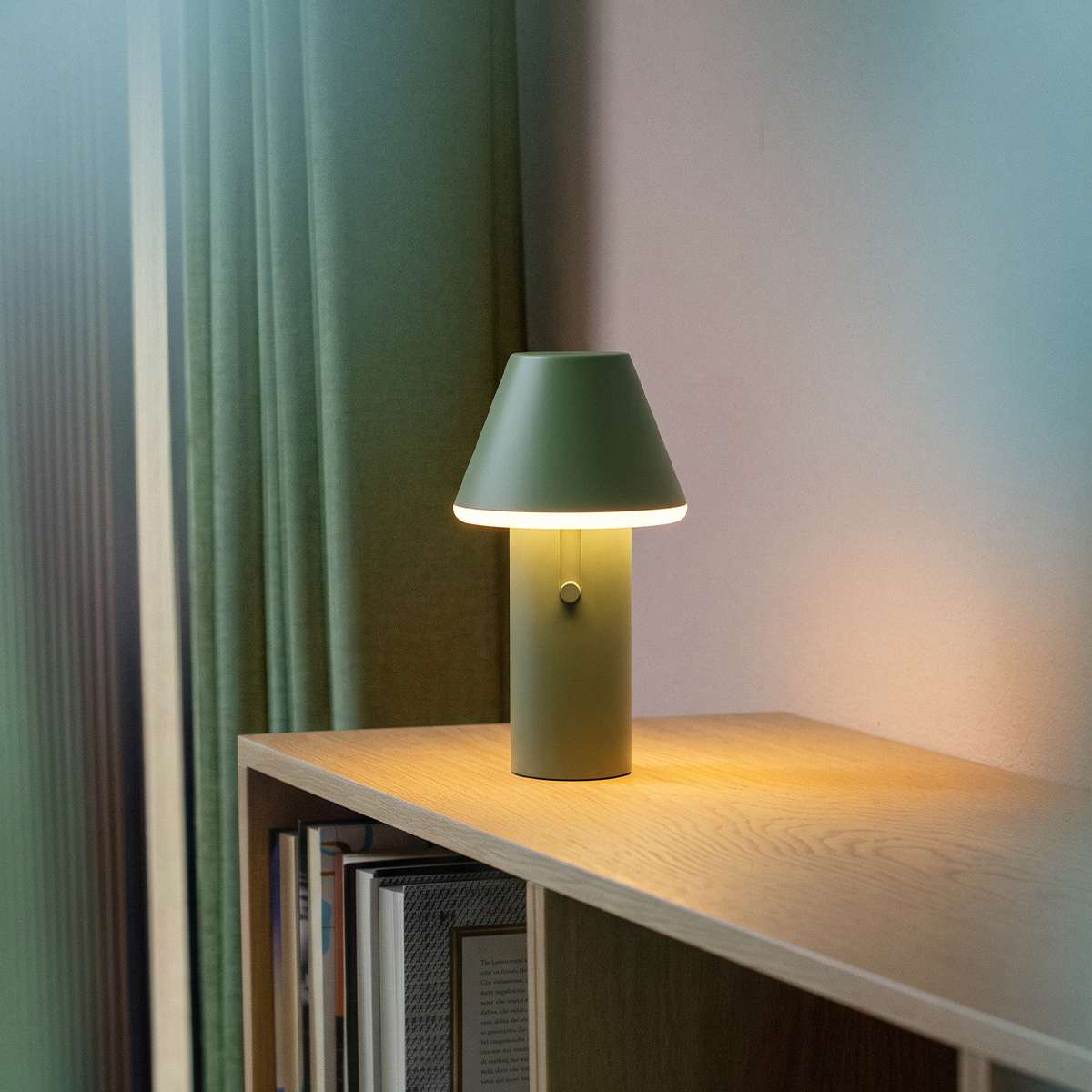sackit Cone cordless table lamp in green glowing softly on a wooden shelf beside books in a cosy indoor setting