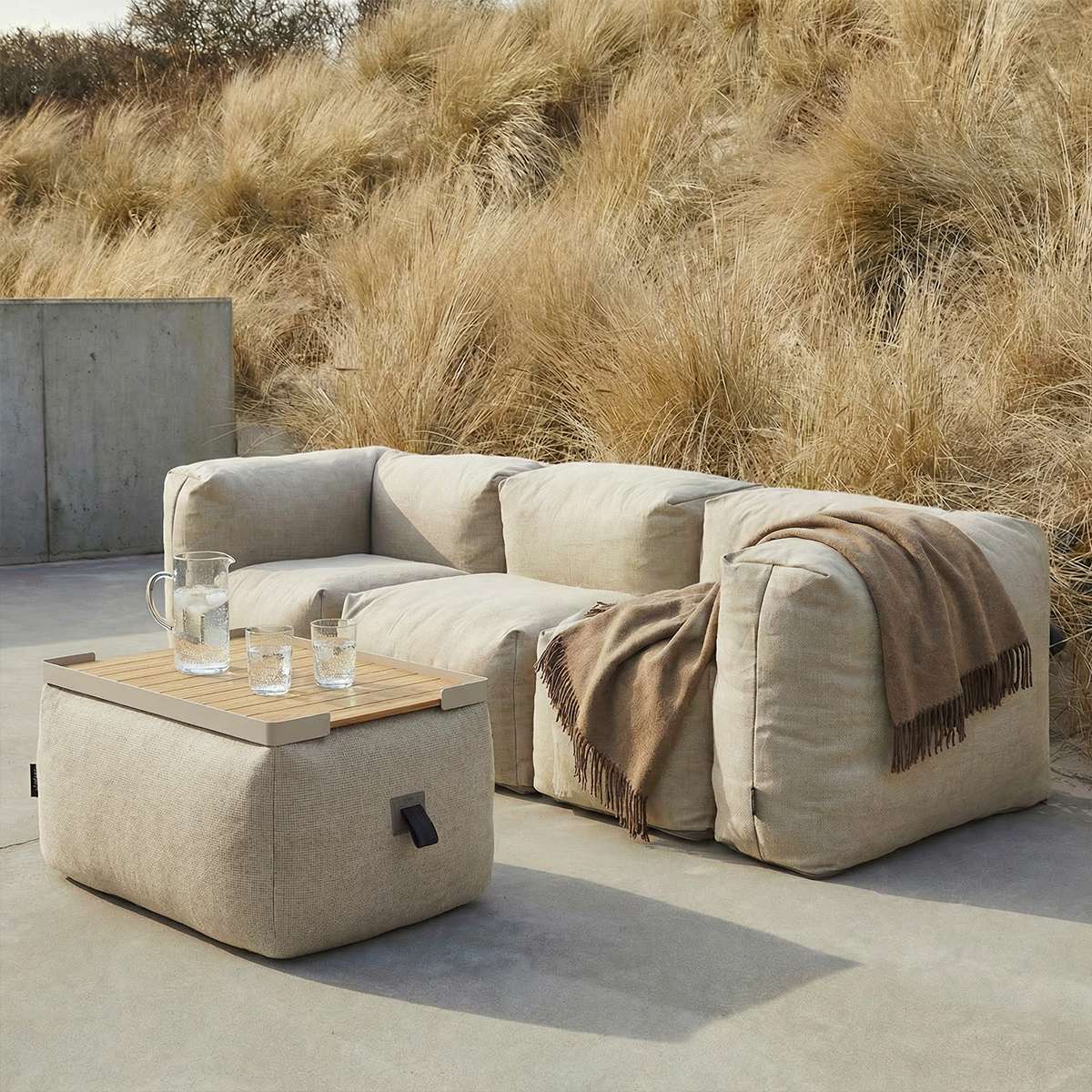 Beige outdoor pouf with a wooden tray beside a matching lounge sofa and blanket in a Nordic garden setting by sackit