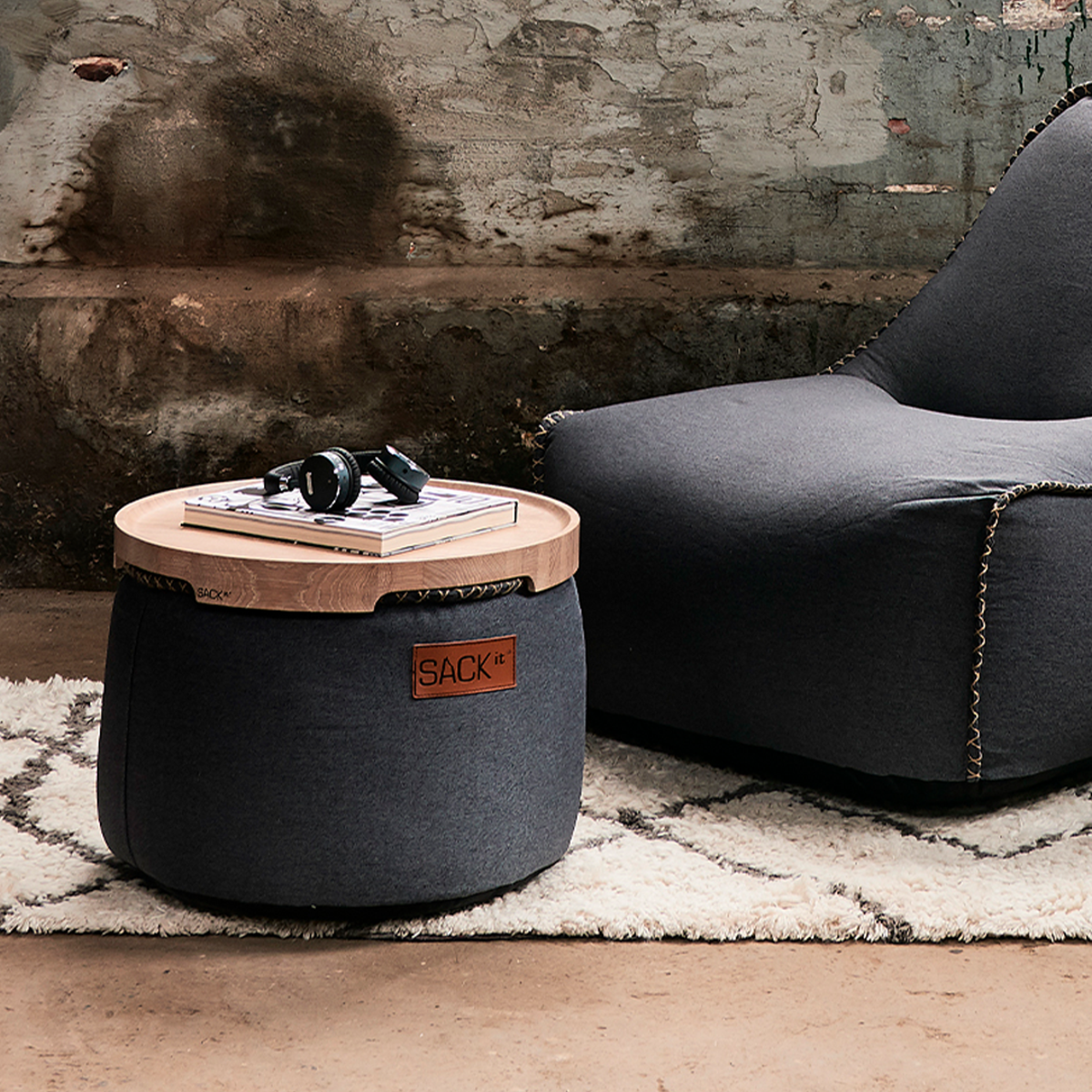 Dark gray sackit canvas pouf with a wooden tray holding books and headphones beside a matching lounge chair in a rustic indoor setting