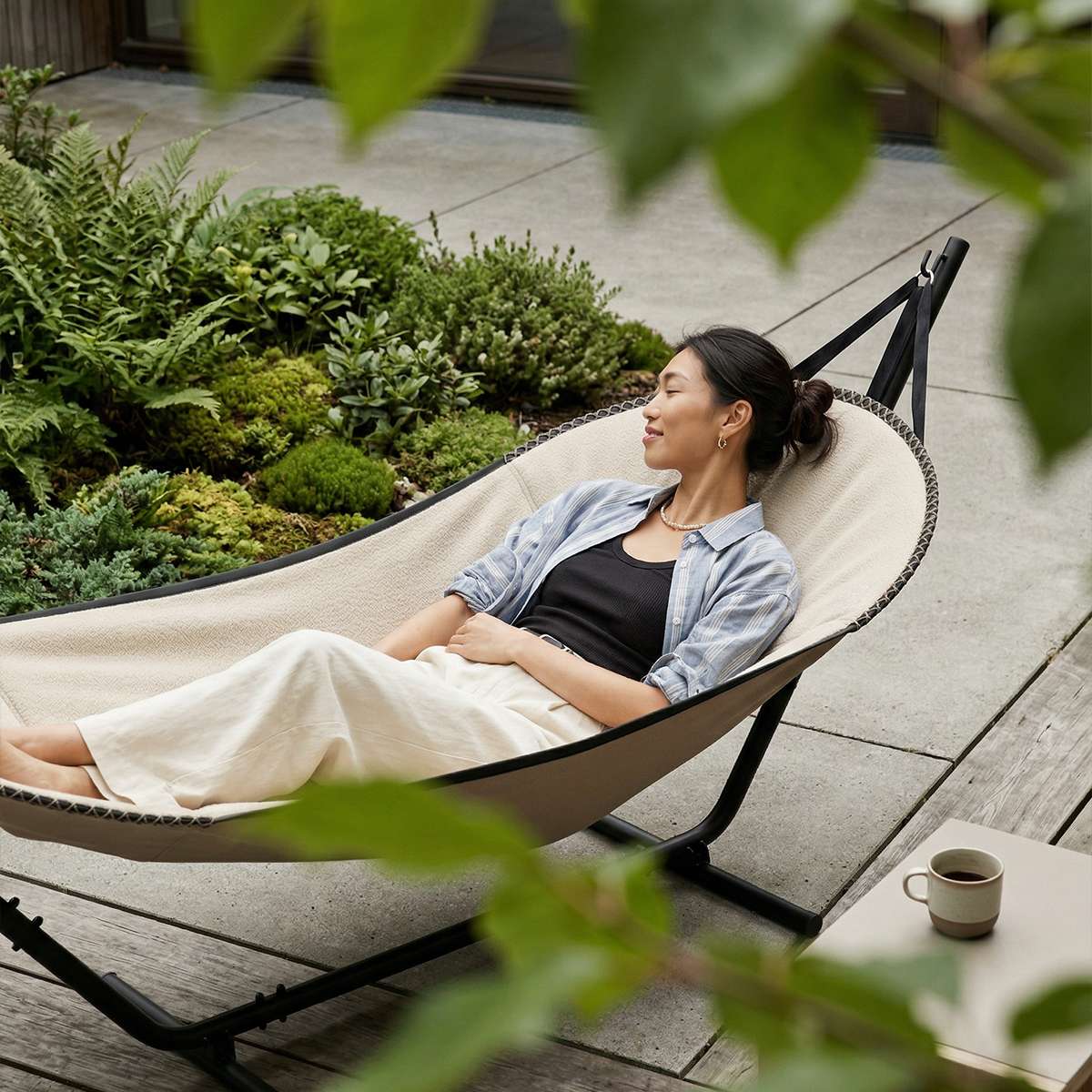Woman relaxing in a Cobana hammock on a garden patio surrounded by lush greenery with a cup of coffee nearby