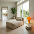 Soft indoor lounge sofa in textured beige upholstery beside floor to ceiling windows in a bright Nordic home with an orange lamp and glass side table by sackit